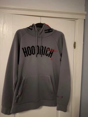 Grå hoodie från Hoodrich  - Snygg grå hoodie från Hoodrich med svart och rött broderat tryck på bröstet. Stor känguruficka med dragkedja, huva med dragsko och tryck även på ryggen. Perfekt för en streetwear-look och riktigt bekväm med mjukt material.
