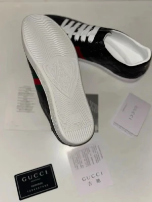 Svarta sneakers med gröna och röda ränder - Säljer ett par svarta sneakers i skinn med präglad logga och klassiska gröna och röda ränder på sidan. Skorna har vit sula och vita skosnören. Perfekta för dig som vill ha en clean men ändå lyxig look.