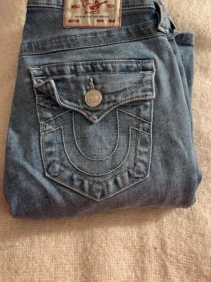 True Religion Jeans - Säljer ett par ljusblå True Religion Becca jeans med bootcut och snygga detaljerade bakfickor. Mid rise modell med klassisk silverknapp och kontrastsömmar. Perfekta för dig som gillar en avslappnad men ändå trendig look.