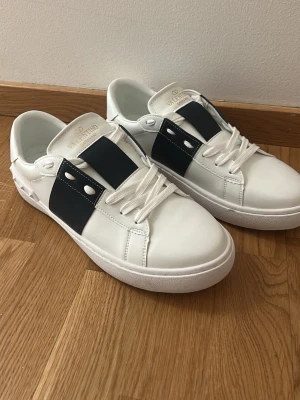 Valentino Garavani vita sneakers - Snygga vita sneakers från Valentino Garavani med bred svart rem och vita nitar på sidorna. Klassisk rund tå, vita snören och platt sula. Tillverkade i skinn för en lyxig känsla och stilren look.