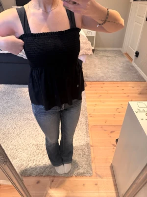 Svart smockad linnetopp - Svart linnetopp med breda axelband och smockad överdel. Toppen har en luftig och lätt utsvängd passform nedtill, perfekt för en avslappnad och trendig look. Tillverkad i mjukt material som känns skönt mot huden. Du kan även använda denna topp som en kjol genom att bara vika in banden. 