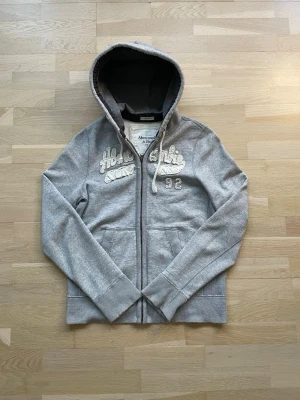 Grå hoodie från Abercrombie & Fitch - Snygg grå hoodie med dragkedja från Abercrombie & Fitch. Broderad text och siffran 92 på bröstet, samt två fickor framtill. Mjuk insida och justerbar huva med snören. Perfekt för en avslappnad och trendig stil.