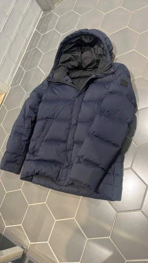 Mörkblå dunjacka från J.Lindeberg - Snygg mörkblå dunjacka från J.Lindeberg med huva och dragkedja. Jackan har en klassisk pufferlook med breda kanaler och diskret logga på ärmen. På vänstra ärmen finns det en liten öppning som sytts ihop och är knappt märkbart.  Perfekt för kalla dagar och riktigt skön att bära under vintern.