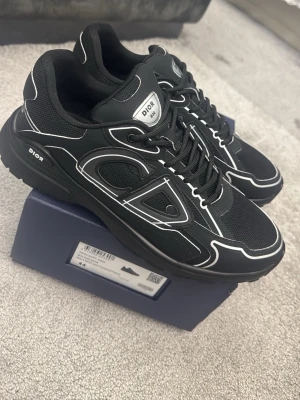 Svarta Dior B30 sneakers - Säljer ett par svarta Dior B30 sneakers med vita detaljer och stora CD-loggor på sidorna. Skorna har snörning, rund tå och är tillverkade i en mix av mesh och syntetmaterial för en sportig och modern look.