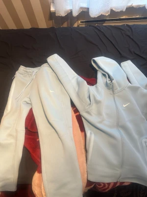 Ljusblåa nocta tech fleece traksuit från Nike - Säljer ett par ljusblåa mjukisbyxor från Nike med dragsko i midjan och diskret Nike-logga på benet. Byxorna har en relaxed passform och är gjorda i mjukt material, perfekta för chill dagar. Matcha med hoodie för en clean look.