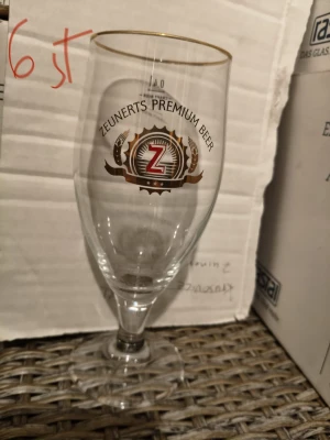 Zeunerts Premium Beer ölglas - Snyggt ölglas från Zeunerts Premium Beer med guldkant upptill och tryckt logga i guld, svart och rött. Glaset har en klassisk ölglasform med fot och kupa, perfekt för att njuta av en kall öl. Tillverkat i klart glas och dekorerat med detaljerad emblem.