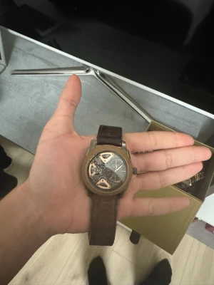 Fossil  klocka med läderband - Snygg klocka med brunt läderband och rund boett i trälook. Urtavlan är mörk med synliga mekaniska detaljer och silverfärgade visare. Klockan har en unik design med öppna partier som visar urverket. Perfekt för dig som gillar stilrena accessoarer med en twist.