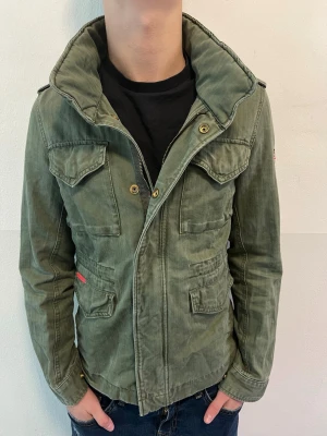 Grön militärinspirerad jacka Superdry - Snygg grön jacka från Superdry med militärvibbar. Jackan har fyra fickor framtill, hög krage och patch med brittiska flaggan på ärmen. Stängs med dragkedja och knappar. Perfekt för dig som gillar streetwear och utilitystil.