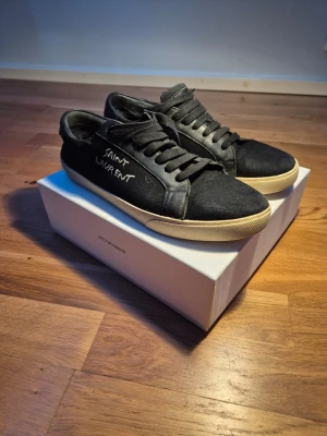 Svarta Saint Laurent sneakers - Svarta sneakers från Saint Laurent med vit broderad logga på sidan. Skorna har snörning, rund tå och platt sula. Ovandelen är i textil och detaljer i skinn, med en beige sula som kontrast. Perfekta för en stilren och trendig look.