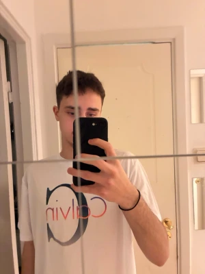 Vit Calvin Klein t-shirt med tryck - Vit t-shirt från Calvin Klein med stort C och Calvin-tryck i svart, oranget/blått på bröstet. Klassisk rund hals och korta ärmar. Perfekt för en stilren och modern look. Inte äkta!! Hör gärna av dig vid frågor eller liknande🙏