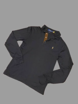 Svart långärmad piké från Polo Ralph Lauren - Snygg svart långärmad pikétröja från Polo Ralph Lauren i slim fit-modell. Tröjan har bruna detaljer vid knäppningen, klassisk krage och den ikoniska broderade loggan på bröstet. Perfekt för en stilren och avslappnad look.