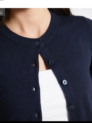 Mörkblå kofta med rund hals - Mörkblå kofta med rund halsringning och knappar framtill. Koftan har långa ärmar och ribbade muddar vid ärmslut och nederkant. Perfekt att slänga över en t-shirt för en enkel och stilren look.