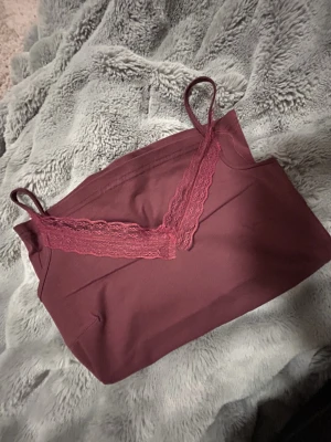 Vinrött linne med spetsdetalj - Mysigt vinrött loungewear-linne med tunna axelband och dekorativ spets längs urringningen. Perfekt för chill hemma eller som lager under en kofta. Mjukt material som känns skönt mot huden.