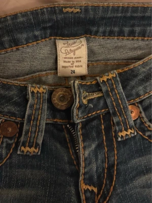 Blå True Religion bootcut jeans - Säljer ett par blå jeans från True Religion med snygga kontrastsömmar och klassisk bootcut-modell. Jeansen har låg midja och är tillverkade i slitstarkt denim med coola detaljer på fickorna. Perfekta för dig som gillar en avslappnad men trendig stil.