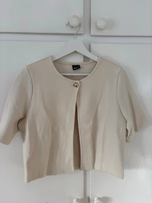 Beige kofta/topp från Gina Tricot - Stilren beige kofta från Gina Tricot med korta ärmar och rund halsringning. Koftan är öppen framtill och stängs med en guldfärgad knapp upptill. Storlek m🩷