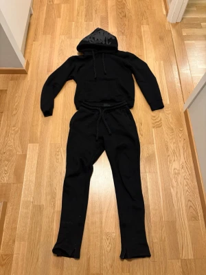 Syna World Set - Säljer ett svart set med hoodie och byxor i mjukt material. Hoodien har dragsko och tryck i grått på huvan, medan byxorna har resår i midjan och matchande tryck baktill. Perfekt för en avslappnad stil.