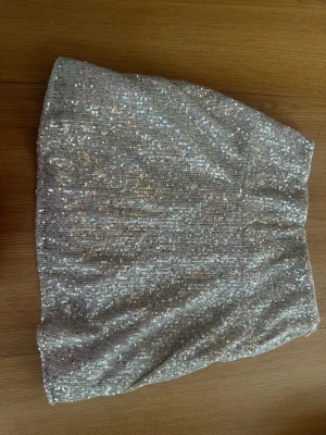Glittrig silver kortkjol - Säljer en glittrig silverfärgad kortkjol med paljetter. Kjolen har en rak passform och är fodrad med ett mjukt vitt tyg på insidan för extra komfort. Perfekt för dig som vill sticka ut och glänsa lite extra.