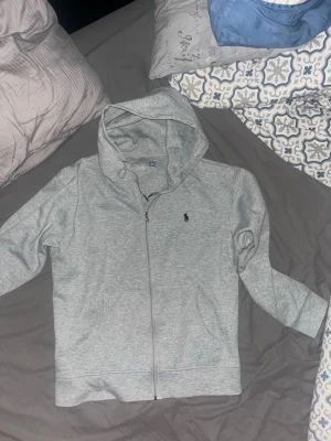 Grå zip hoodie från Polo Ralph Lauren - Snygg ljusgrå hoodie med dragkedja från Polo Ralph Lauren. Klassisk huva, två fickor framtill och den ikoniska mörkblå loggan broderad på bröstet. Perfekt till chill eller streetwear-stil. Mjuk bomullskänsla och relaxed fit. Inga kändare skador på tröjan och den är relativt oanvänd 