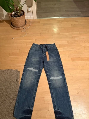Blå raka jeans med slitningar - Säljer ett par blå raka jeans från Dsquared2 med coola slitningar och patchdetaljer på baksidan. Jeansen har klassisk femficksdesign och en avslappnad passform. Perfekta för dig som gillar streetstyle och vill sticka ut lite extra.
