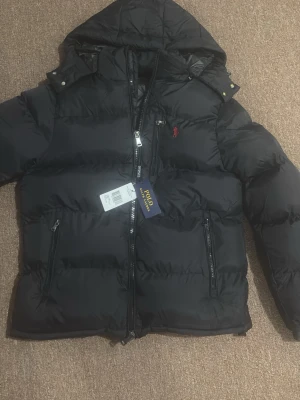 Svart dunjacka från Polo Ralph Lauren - Svart puffer dunjacka från Polo Ralph Lauren med huva, dragkedja framtill och flera fickor med dragkedja. Jackan har det klassiska röda Polo-logotypen broderad på bröstet och en quiltad design som ger en schysst streetstil.