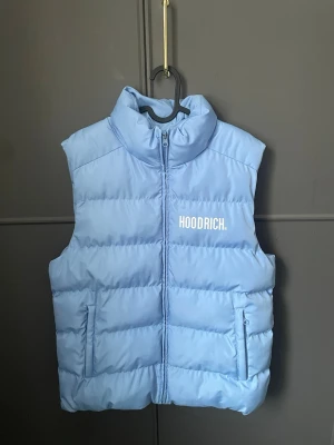 Ljusblå dunväst från Hoodrich - Snygg ljusblå dunväst från Hoodrich med hög krage och dragkedja framtill. Västen har två fickor med dragkedja och en stilren logga på bröstet. Perfekt för lager på lager och streetwear-vibe.