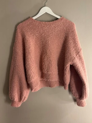 Rosa fluffig stickad tröja Jolio&Co - Mysig rosa stickad tröja från Jolio&Co med fluffig textur och avslappnad passform. Tröjan har ballongärmar och ribbade muddar vid ärmslut och nederkant. Perfekt för lager på lager och ger en trendig look till din outfit.