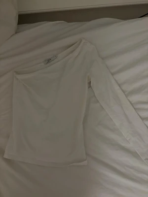 Vit oneshoulder topp från NA-KD - Säljer en stilren vit oneshoulder topp från NA-KD. Toppen har lång ärm på ena sidan och ärmlös på andra, vilket ger en trendig asymmetrisk look. Tillverkad i mjukt material som känns skönt mot huden. Perfekt för dig som vill sticka ut lite extra.