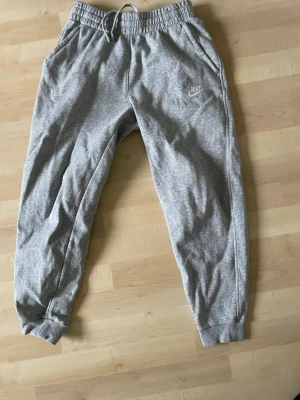 Gråa mjukisbyxor från Nike - Snygga gråa mjukisbyxor från Nike med broderad logga på vänster ben. Byxorna har resår i midjan med snörning och muddar vid bensluten. Tillverkade i mjukt bomullsmaterial för maximal komfort. Perfekta för chill eller träning. Säljer för att de är för små för mig. Pris är inte hugget i sten!