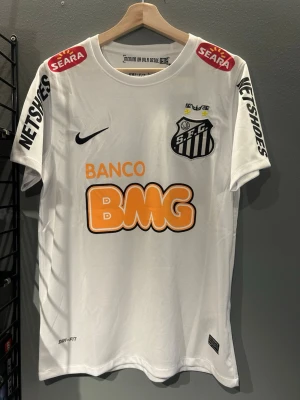 Santos FC Neymar Jr matchtröja - Vit Santos FC matchtröja från Nike med Neymar Jr och nummer 11 på ryggen. Tröjan har svarta och orangea tryck, klubbmärke på bröstet och flera sponsorer. Tillverkad i Dri-Fit material som andas, perfekt för fotbollsträning eller samling. Helt ny med tagg en replika