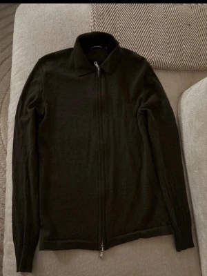JL Mörkgrön stickad ziptröja med krage - Säljer en mörkgrön stickad tröja med dragkedja framtill och klassisk krage. Tröjan har långa ärmar och är tillverkad i ett mjukt, tunt stickat material. Perfekt för lager på lager och enkel att matcha med olika outfits.