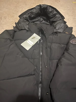 Svart dunjacka från Canada Goose XS - Snygg svart dunjacka från Canada Goose med hög krage och huva. Jackan har dragkedja och knappar framtill samt en patch med logga på ärmen. Perfekt för kalla vinterdagar och riktigt stilren look.