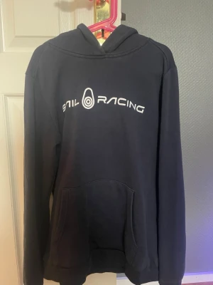 Marinblå hoodie från Sail Racing - Säljer en marinblå hoodie från Sail Racing med vit logga tryckt på bröstet. Tillverkad i mjukt och bekvämt material som passar perfekt för en avslappnad stil.