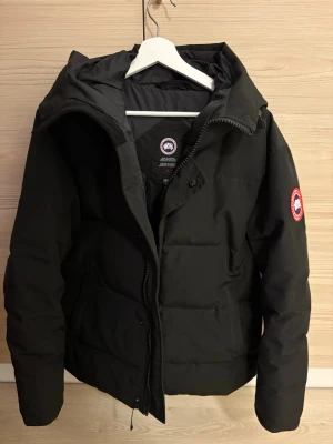 Canada Goose MacMillan - Säljer en svart pufferjacka från Canada Goose med klassisk logga på ärmen. Jackan har hög krage, dragkedja framtill och är vadderad för extra värme. Perfekt för kalla dagar och har en stilren look som funkar till det mesta.