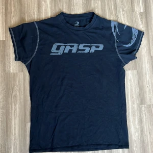 Svart GASP t-shirt med tryck - Svart t-shirt från GASP i bomull med grått tryck över bröstet och detaljer på ena ärmen. Klassisk rund halsringning och kontrastsömmar vid axlarna. Perfekt för dig som gillar sportig och avslappnad stil.