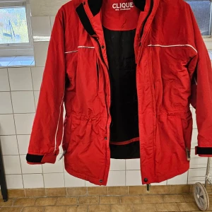 Röd vindjacka från Clique med huva - Röd vindjacka från Clique med reflekterande detaljer, justerbar huva och dragkedja framtill. Jackan har elastisk midja och muddar med kardborre vid ärmslut. Perfekt för blåsiga dagar och enkel att matcha med streetwear.