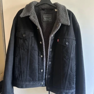 Svart teddyfodrad jeansjacka Levi's - Svart jeansjacka från Levi's med grått teddyfoder och krage. Klassisk modell med knappar framtill, två bröstfickor och sidofickor. Perfekt för kyliga dagar och har en snygg, tidlös look med en loose passform.