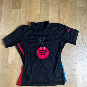 FC Barcelona svart matchtröja Nike - Snygg svart FC Barcelona fotbollströja från Nike med röda och blå detaljer, Spotify-logga på bröstet och Lamine Yamal 19 på ryggen. Tröjan har klubbmärke på bröstet, korta ärmar och är tillverkad i lätt funktionsmaterial.