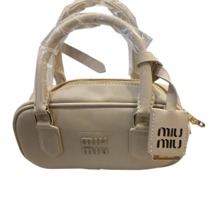 Beige handväska  - Snygg beige handväska  med stilren, rektangulär form och präglad logga framtill. Väskan har dubbla handtag, guldfärgade detaljer och en praktisk dragkedja upptill. Tillverkad i slätt skinn med matchande tag med logotyp.