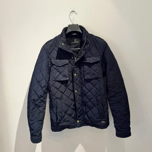 Mörkblå quiltad jacka Scotch & Soda - Quiltad mörkblå jacka från Scotch & Soda med guldiga knappar och två stora bröstfickor. Jackan har klassisk krage, lång ärm och snyggt rutmönster. Perfekt för höst och vår och ger dig en stilren look. En knapp saknas men det är inget man tänker på, hör av er vid frågor 