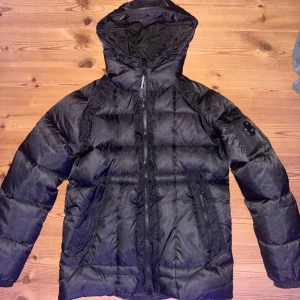 CP jacka - Svart pufferjacka från Cp company med huva och dragkedja framtill. Jackan har en quiltad design, två sidofickor med dragkedja och en logotyp på ärmen. Strl M men skulle nog passa S med. Bara att höra av dig vid frågor!