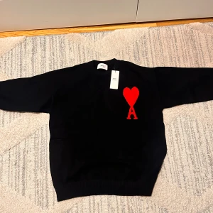 Svart stickad tröja från Ami Paris - Svart stickad tröja från Ami Paris med rund halsringning och ikoniskt rött hjärta med bokstaven A broderat på bröstet. Tröjan har ribbade muddar och är långärmad, perfekt för en stilren och trendig look.
