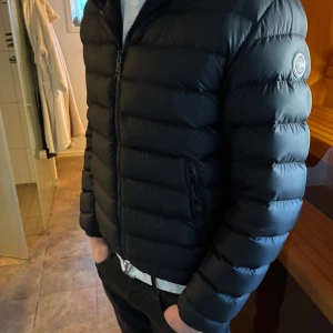 Svart Colmar pufferjacka herr - Säljer en svart pufferjacka från Colmar med klassisk rund logotyp på ärmen. Jackan har dragkedja framtill, två fickor med dragkedja och en stilren, quiltad design. Perfekt för kalla dagar och ger en clean look till din outfit. Jackan har hål på ärmarna, går lätt att laga men är för liten så har inte lagat det själv. Den är i storlek M/L              Köpt på boozt för 3700kr på rea.