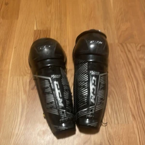 Svarta benskydd CCM Tacks 10" - Säljer ett par svarta CCM Tacks benskydd med coolt randigt mönster och tydlig logga. De har justerbara remmar med gul detalj och är gjorda i slitstark syntet och plast. Perfekta för hockey eller annan sport där du behöver skydda benen.