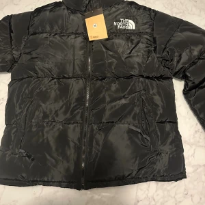 Svart pufferjacka från The North Face - Säljer en svart pufferjacka från The North Face med klassisk logga på bröstet och ryggen. Jackan har dragkedja framtill, två sidofickor med dragkedja och hög krage. Materialet är glansigt och känns riktigt mjukt och skönt.