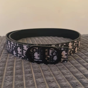 Svart och beige bälte - Snyggt bälte  med klassiskt monogrammönster i svart och beige. Bältet har en stilren, svart spänne med rundade former och är tillverkat i skinn med textil på ovansidan. Perfekt accessoar för att lyfta din outfit.