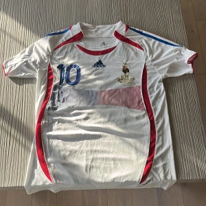 Frankrike Zidane VM 2006 tröja Adidas - Säljer en officiell Frankrike fotbollströja från VM-finalen 2006 med Zidane 10 på ryggen. Tröjan är vit med blå och röda detaljer, broderat FFF-emblem och guldtext. Tillverkad i återvunnen polyester, kortärmad och med klassisk Adidas-design.