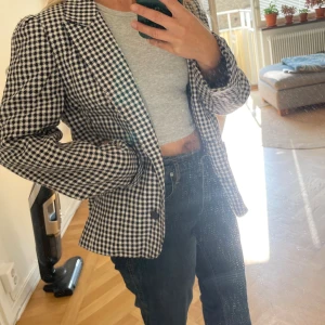 Rutig svartvit kavaj i ullmix - Snygg svartvit rutig kavaj med klassiskt snitt och två knappar framtill. Jackan är fodrad och tillverkad i en ull- och polyestermix, vilket ger en skön känsla och stilren look. Perfekt för dig som vill sticka ut med ett tidlöst mönster.