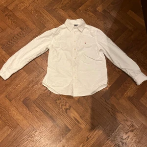 Vit skjorta från Polo Ralph Lauren - Klassisk vit skjorta från Polo Ralph Lauren med broderad logga på bröstet. Skjortan har lång ärm, knappar framtill och button-down krage. Tillverkad i mjuk bomull som känns skön mot huden. Perfekt för en clean och stilren look.