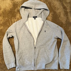 Grå hoodie från Polo Ralph Lauren - Grå hoodie från Polo Ralph Lauren med dragkedja och klassisk broderad logga på bröstet. Tröjan har huva, fickor framtill och är tillverkad i mjuk bomullsmix. Perfekt för chill dagar eller när du vill ha en clean och enkel stil.