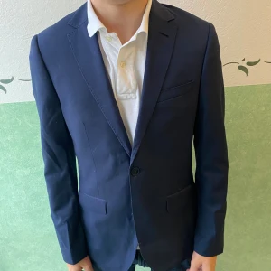 Mörkblå kavaj från Riley Shelby - Snygg mörkblå kavaj från Riley Shelby med klassisk passform. Kavajen har två knappar framtill, tre fickor och ljusblått innerfoder. Perfekt för dig som vill ha en stilren och tidlös look. Materialet känns mjukt och följsamt.